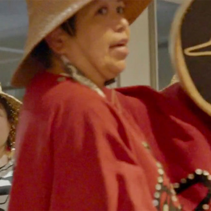 Person in traditional attire, wearing a wide-brimmed hat and holding an item, stands indoors with another similarly dressed person partially visible behind.