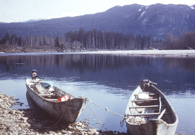 Two small motorboats are docked along a rocky shore, with a calm lake and forested, mountainous backdrop.