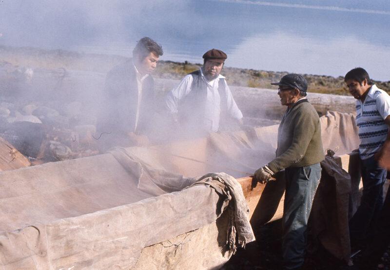 Four men stand around a large, steaming wooden container near a lake, enveloped in mist. One man is lifting a cloth cover.