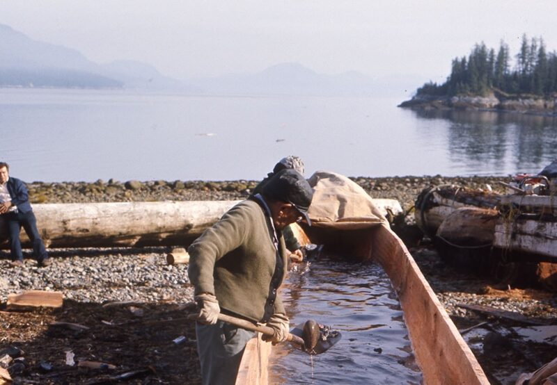 A person carves a wooden canoe near a rocky shore, with a calm lake and forested mountains in the background. Another person stands nearby.