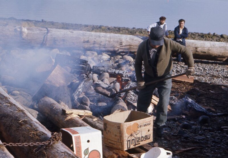 A person tends a fire on a rocky beach surrounded by logs, with two people standing in the background near the shoreline.