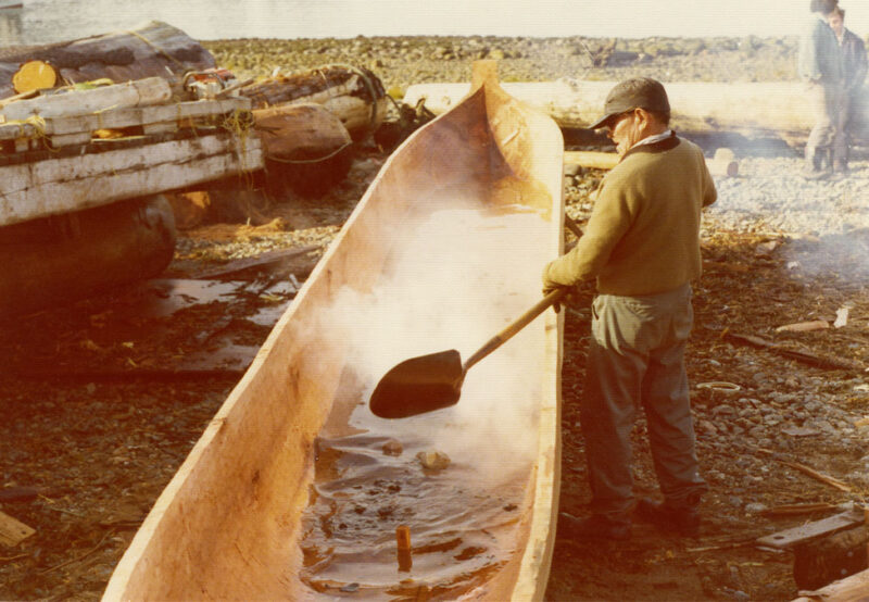 A person uses a paddle to work on a large, hollowed-out wooden canoe near a body of water, surrounded by logs and tools.