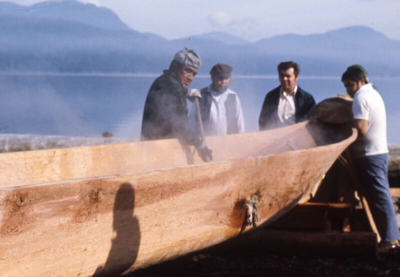 Four people work on carving a large wooden canoe by a misty lakeside, with mountains visible in the background.