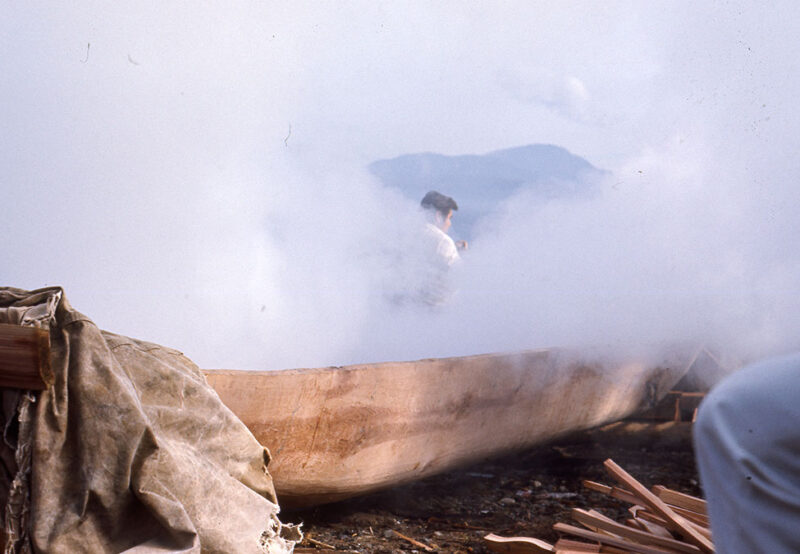 A person surrounded by smoke is seen working on a wooden canoe outdoors, with mountains visible in the background.
