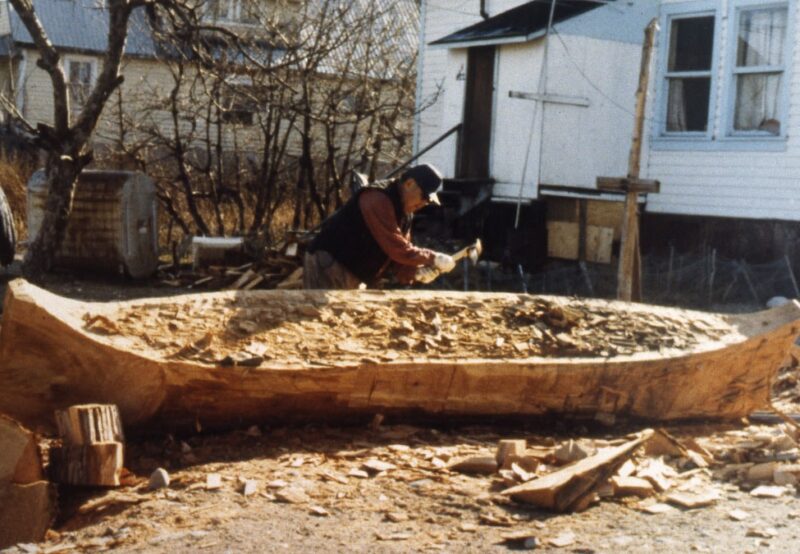 A person carving a large wooden canoe with a hand tool outside near a white house and leafless trees.
