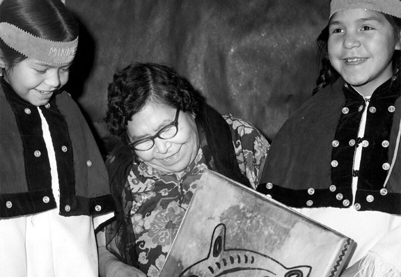 An elderly woman sits between two children wearing traditional attire, all smiling and looking at a piece of art.