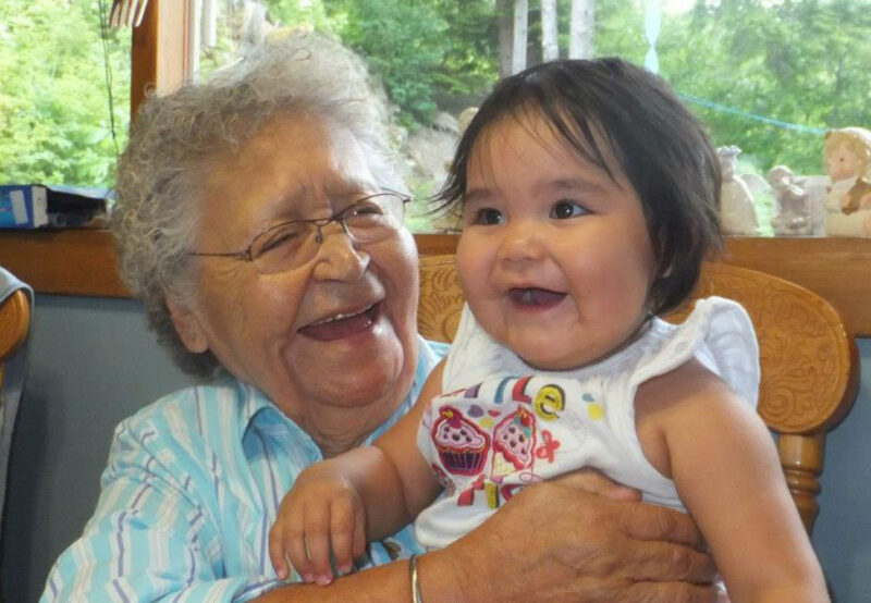 An elderly woman wearing glasses joyfully holding a smiling baby girl on her lap, both in a cozy indoor setting with greenery visible outside.