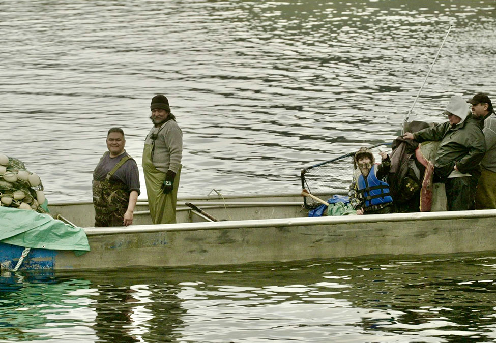 Four people in a small boat on a calm body of water, with fishing gear and supplies onboard. Two are seated while two stand and smile at the camera.