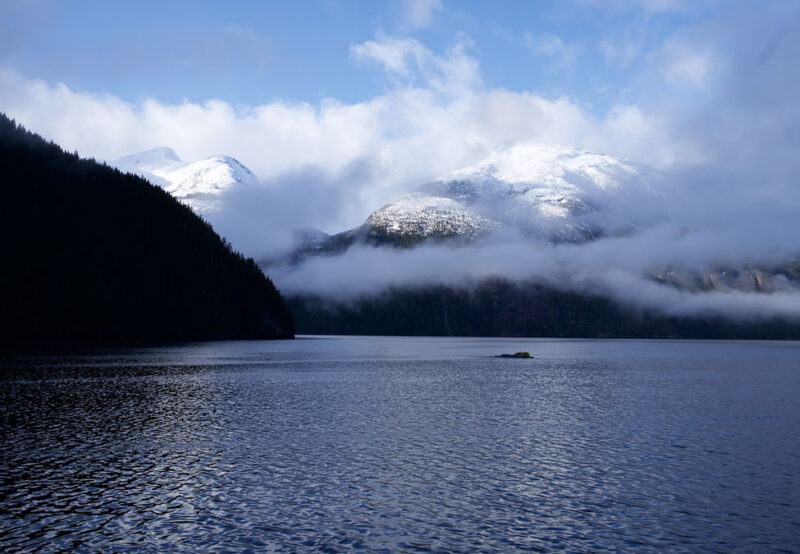 Calm lake with a small rock in the center, surrounded by snowy mountains and mist, under a partly cloudy blue sky.