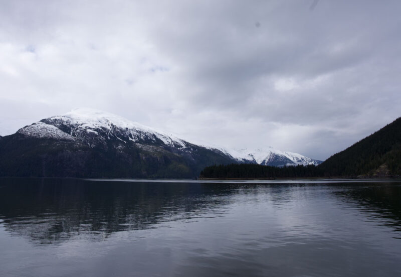 Calm lake with a view of snow-capped mountains and forested hills under a cloudy sky.