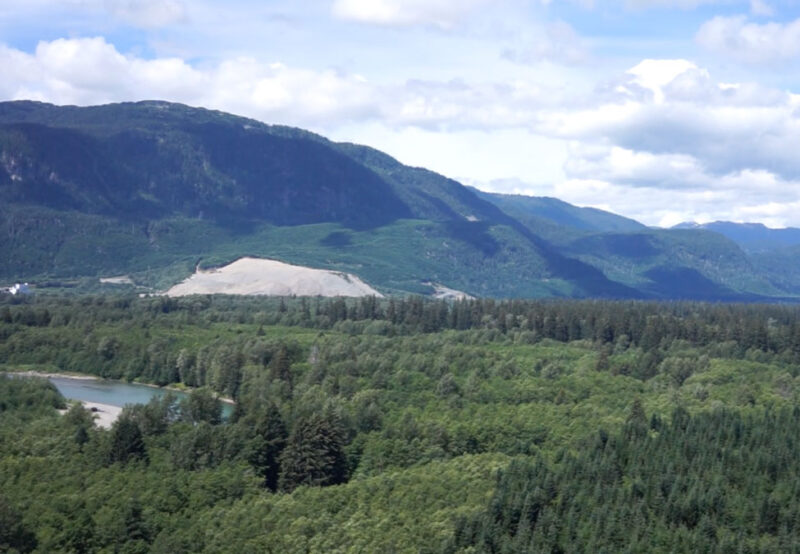 A lush green forest with a river flowing through it, framed by mountainous terrain under a partly cloudy sky.