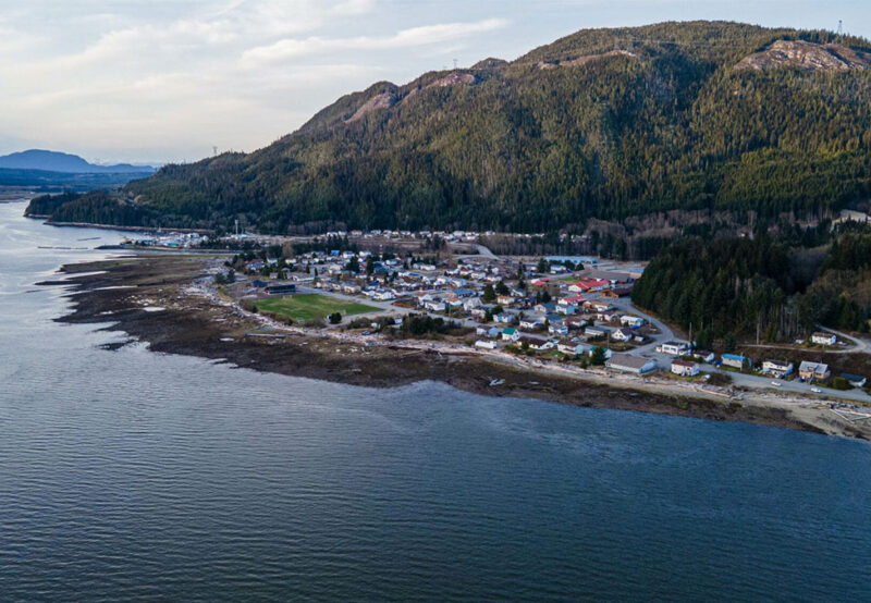 A coastal town at the base of a forested mountain. There are houses, open fields, and a shoreline with a calm body of water.