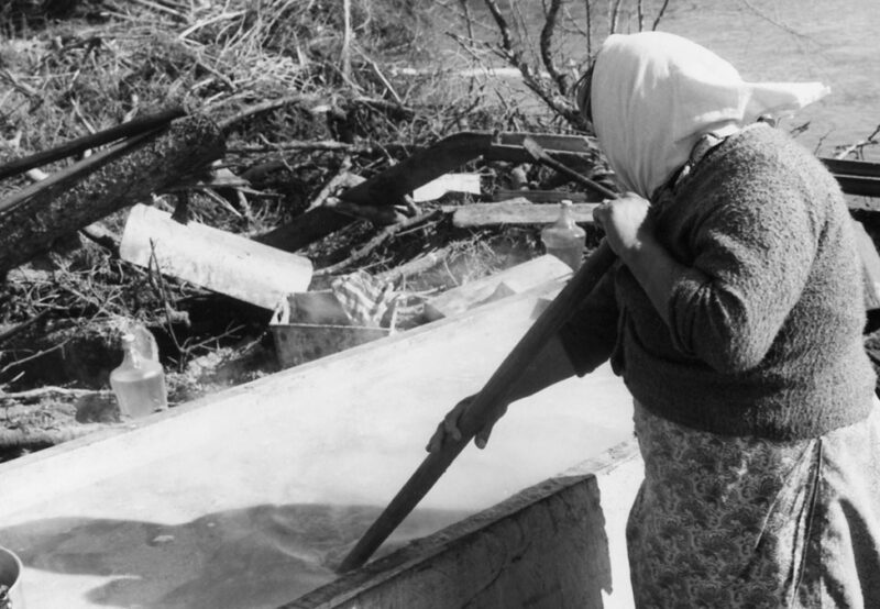 A person wearing a headscarf and sweater stirs soap in a large outdoor basin, surrounded by logs and other debris. The scene is near a river.