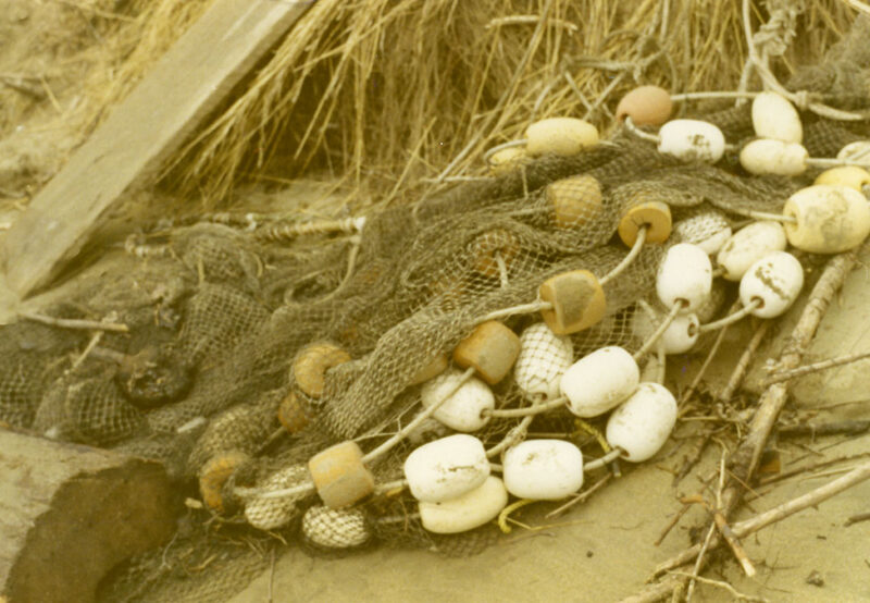Fishing net with white and brown floats tangled and lying on a sandy beach near dried grasses and wooden planks.
