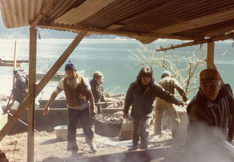 A group of people stands under a rustic shelter near a body of water, carrying fish and engaging in outdoor activities.