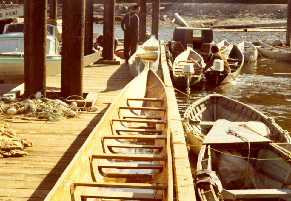 Wooden fishing boats docked at a wooden pier, with one person standing and looking at the boats. Fishing nets are seen on the pier.