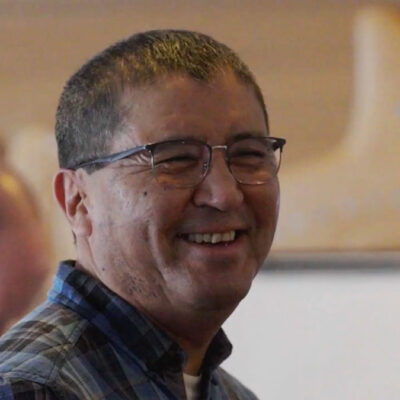 A smiling man in glasses and a checkered shirt stands indoors with a blurred background.