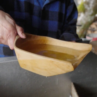 A person in a blue and black plaid shirt holding a wooden bowl containing yellow liquid.