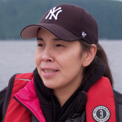 Person wearing a New York Yankees cap and a red life jacket, standing outdoors near a body of water with a forested background.